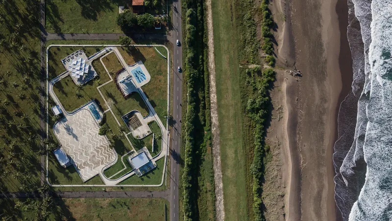 Aerial view of compound with white volumes and serpentine paving adjacent to coastal road and beach