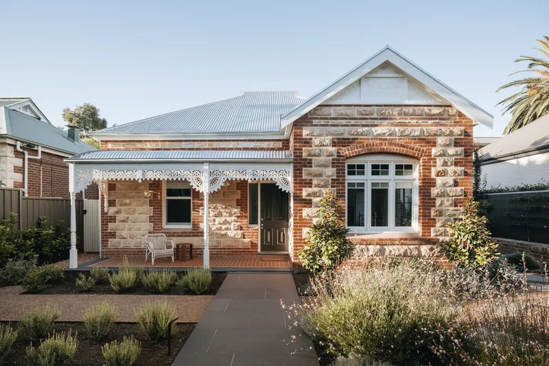 Street facade showing mixed red brick and sandstone walls with planted garden beds and palm tree