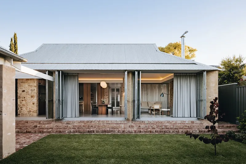 Rear elevation with corrugated metal roof and folding glass doors opening onto brick steps and lawn