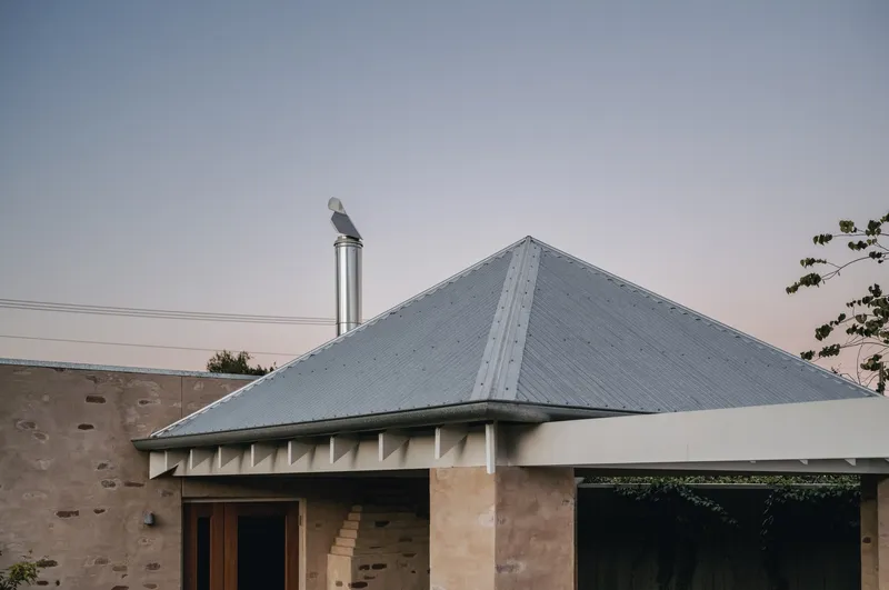 Hipped corrugated metal roof with chimney flue against a pale evening sky