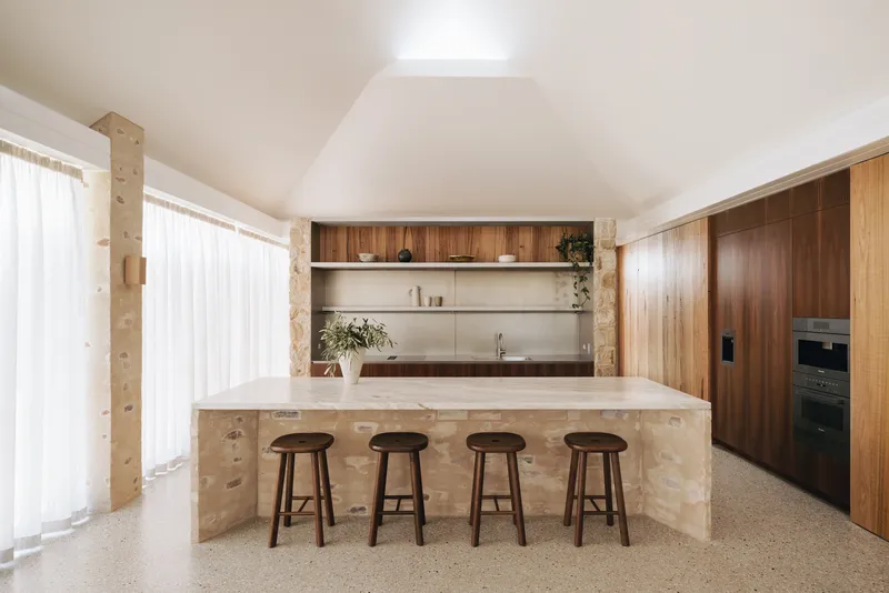 Kitchen island clad in sandstone blocks with four timber stools beneath a vaulted white ceiling