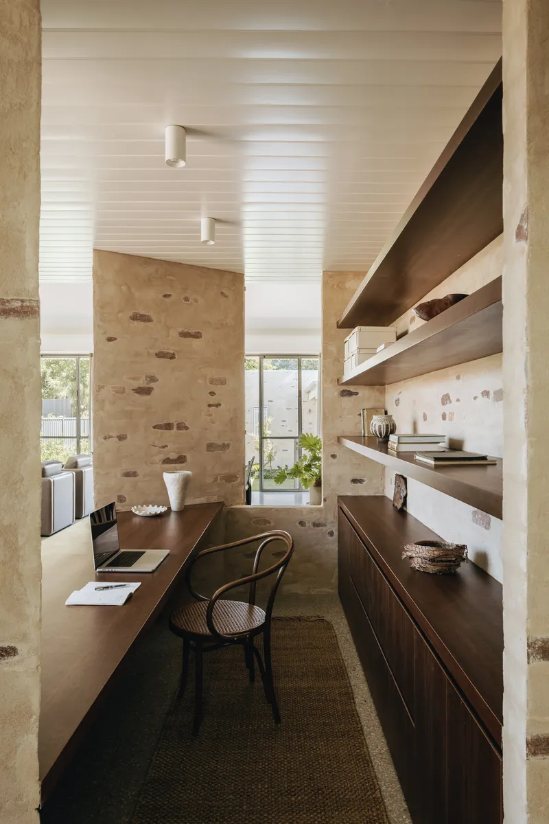 Narrow workspace with exposed stone walls, horizontal wood shelving, white slatted ceiling, and timber desk