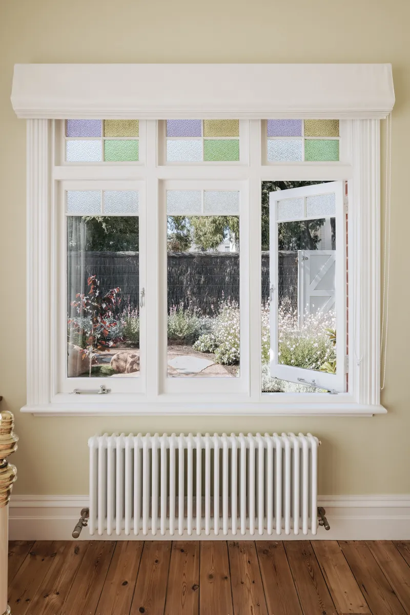White timber window with colored glass transom panels overlooking a garden with flowering plants