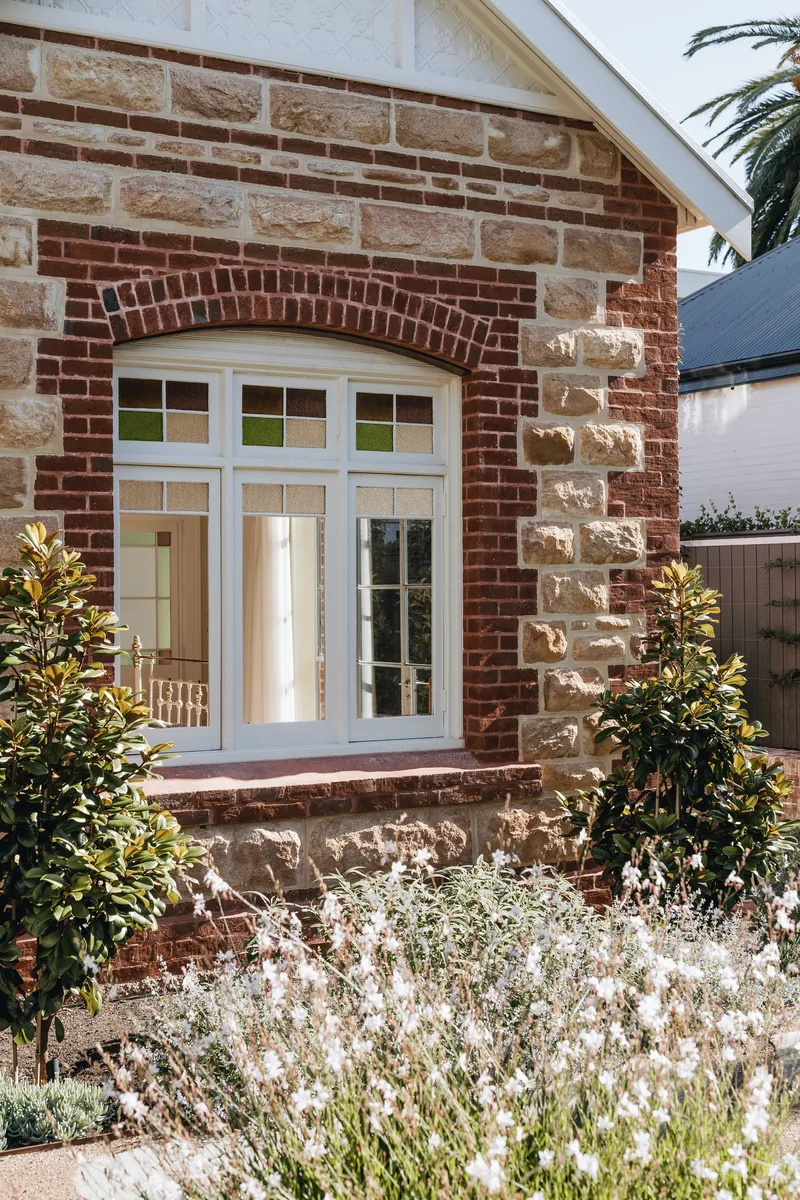 Mixed brick and sandstone facade with arched window above planted beds in afternoon sun