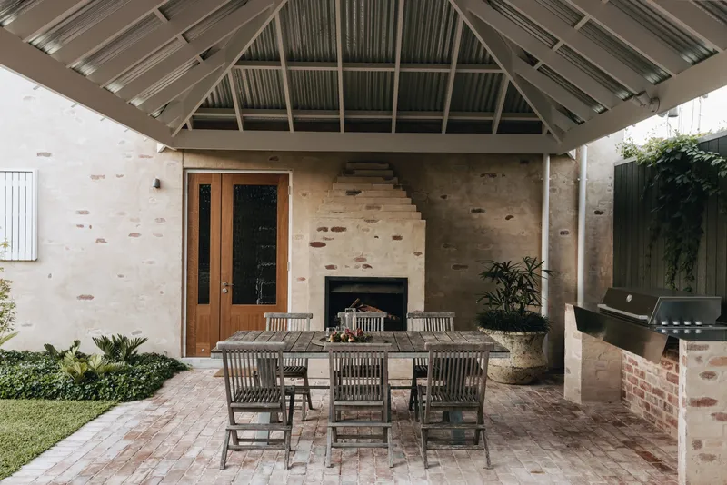 Covered outdoor dining area with exposed timber roof structure and sandstone fireplace wall with brick patches