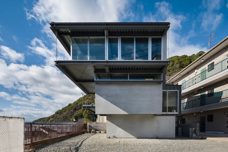 Stacked concrete volumes with cantilevered glass upper floor and gravel courtyard under a partly cloudy sky