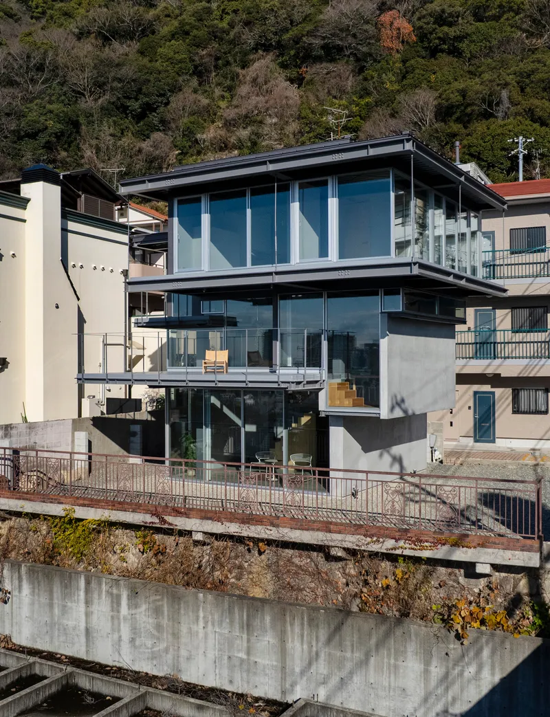 Three-story glass and concrete facade stacked on a hillside retaining wall with neighboring houses and wooded slope behind