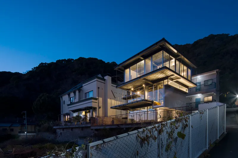 Illuminated steel-framed volume rising above adjacent houses at dusk with forested hillside behind
