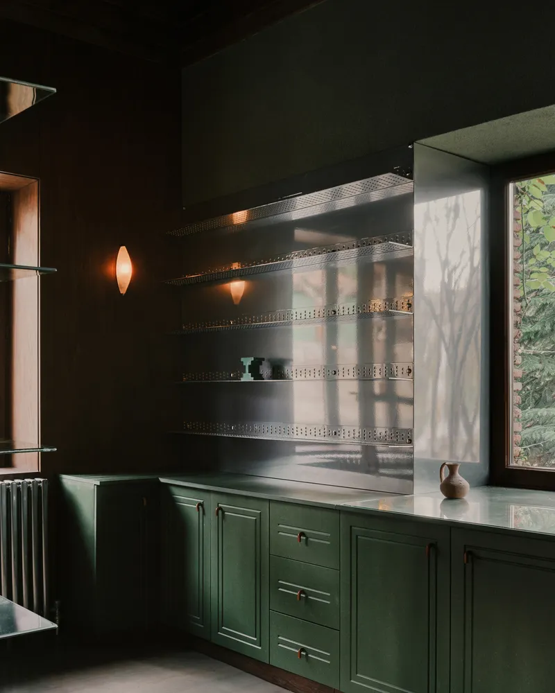 Kitchen alcove with green cabinetry, perforated metal shelves, and sunlight filtering through a window with foliage outside