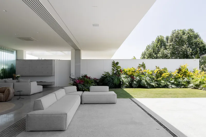 Open-air living area with white sofas on concrete flooring beside a planted garden bed