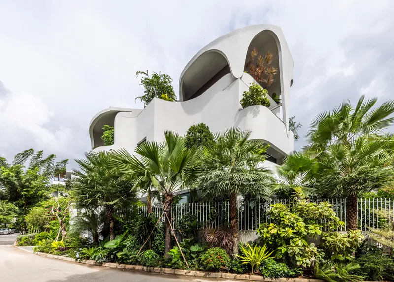 White curving facade with arched openings and rooftop plantings above a planted garden with palms