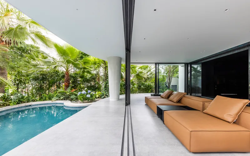 Open living pavilion with tan sectional sofa overlooking a pool and planted garden beds