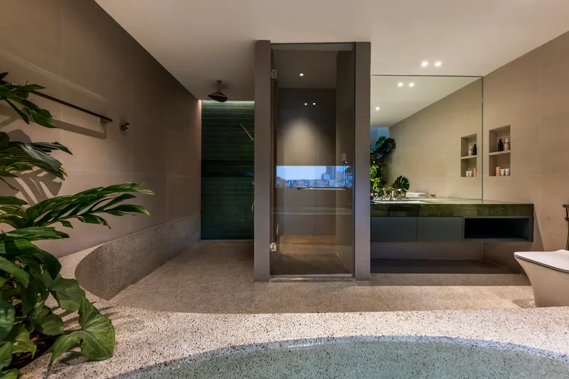 Glass shower enclosure and curved bathtub within a planted bathroom at dusk