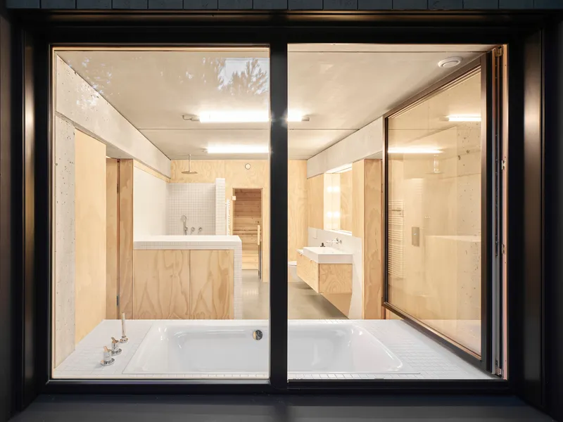 Night view through black metal window frames into bathroom with white tub and plywood cabinetry