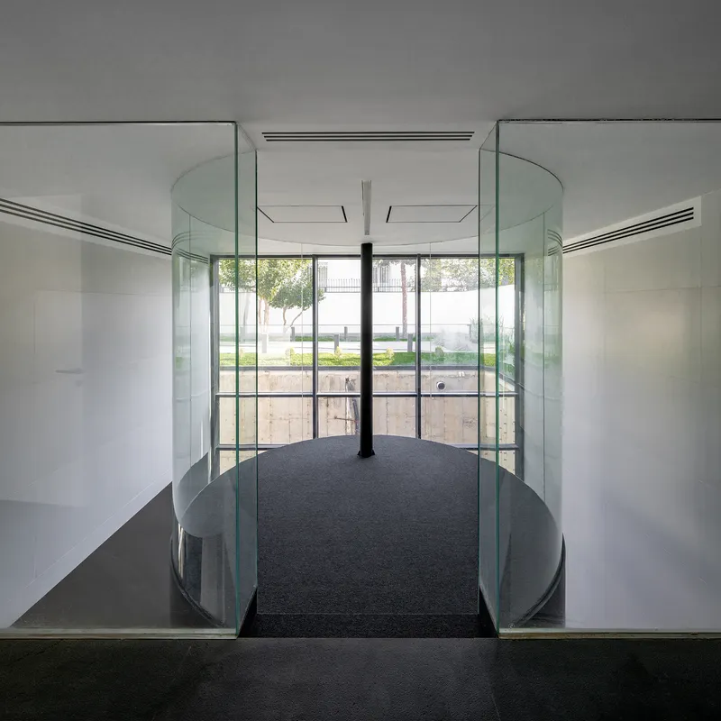Glass-walled bridge with curved railings and central column overlooking a terraced garden courtyard