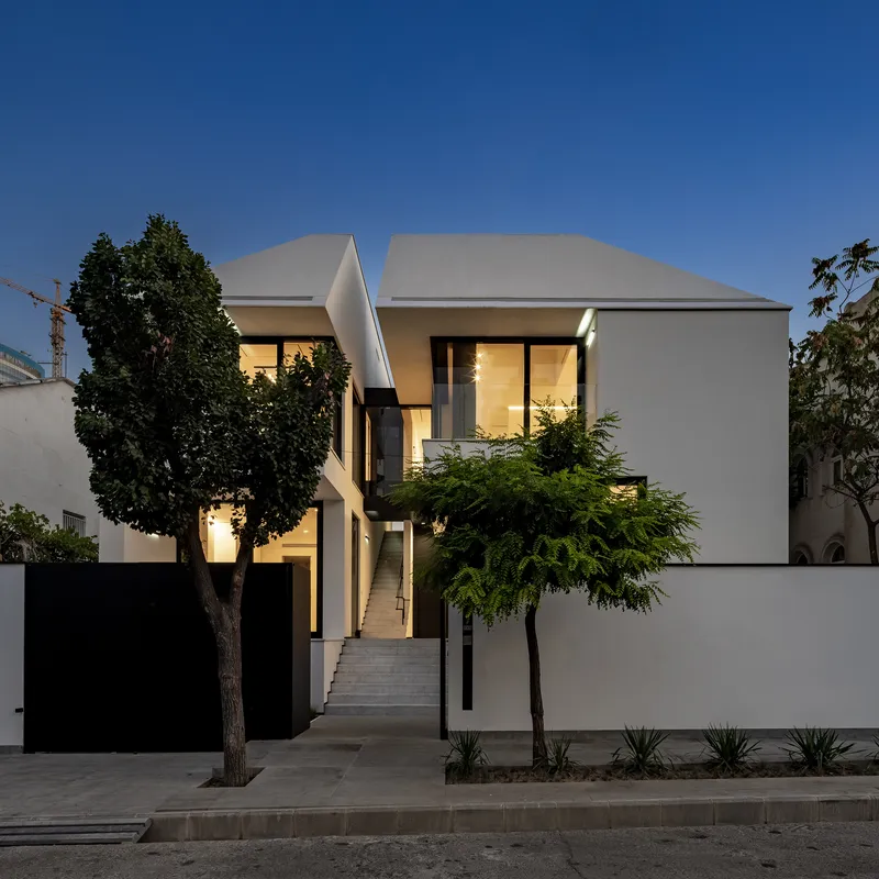 Street view of white stucco facades with illuminated windows at dusk flanked by mature trees