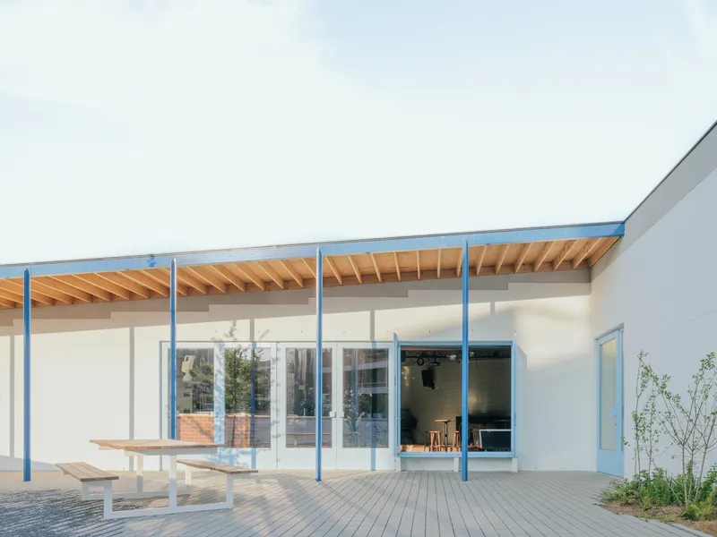 Covered terrace with exposed timber rafters and blue steel columns facing a paved courtyard and planted beds