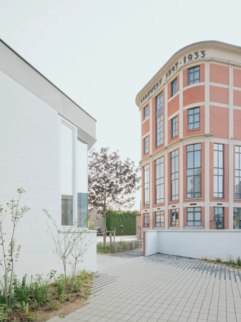 Paved courtyard between white volume and curved brick facade with young trees