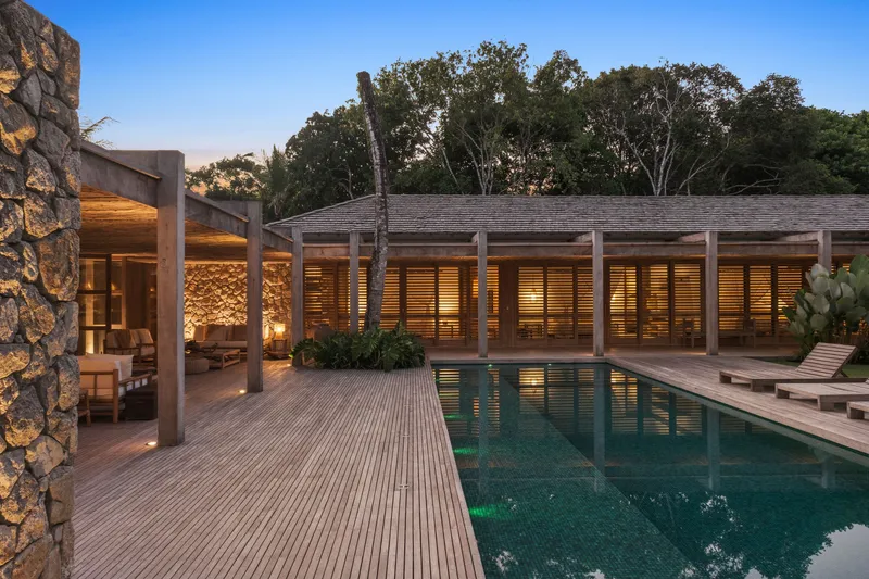 Pool terrace with timber decking and louvered facade wall illuminated at dusk under trees
