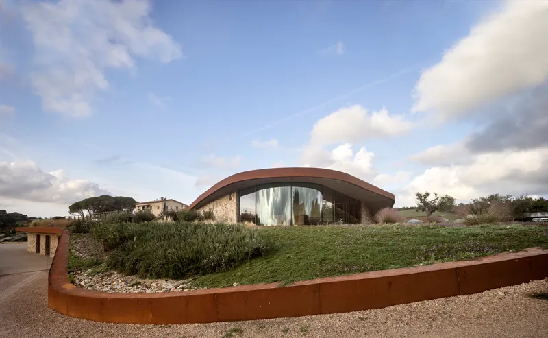 Curving glass-fronted pavilion with planted roof and weathered steel retaining wall under afternoon clouds