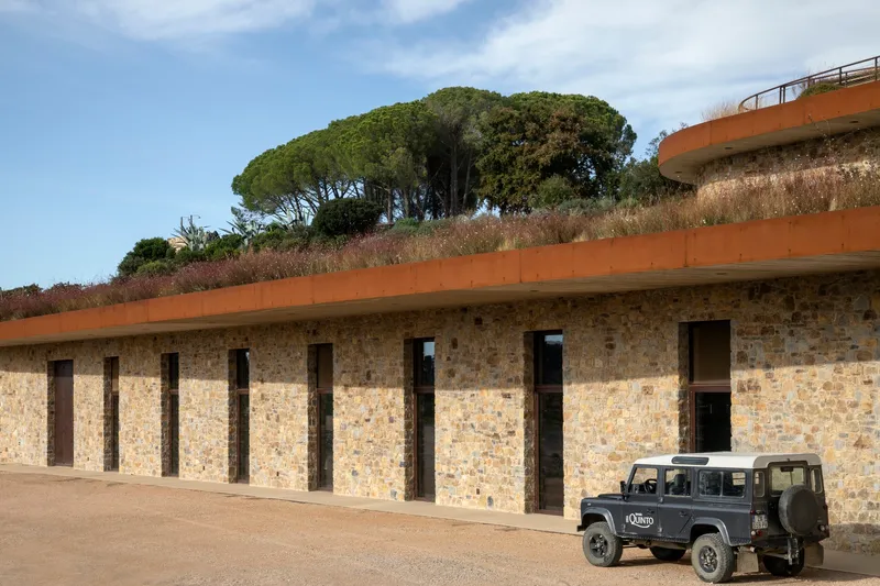 Stone facade with rhythmic timber-framed openings and planted roof edge with parked vehicle below