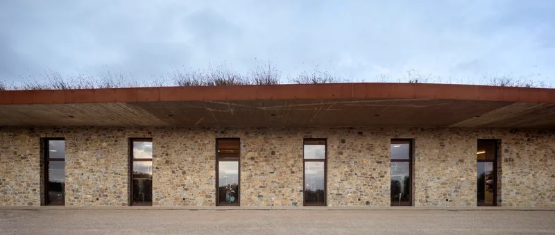 Stone facade with regularly spaced windows and timber door beneath overhanging planted roof