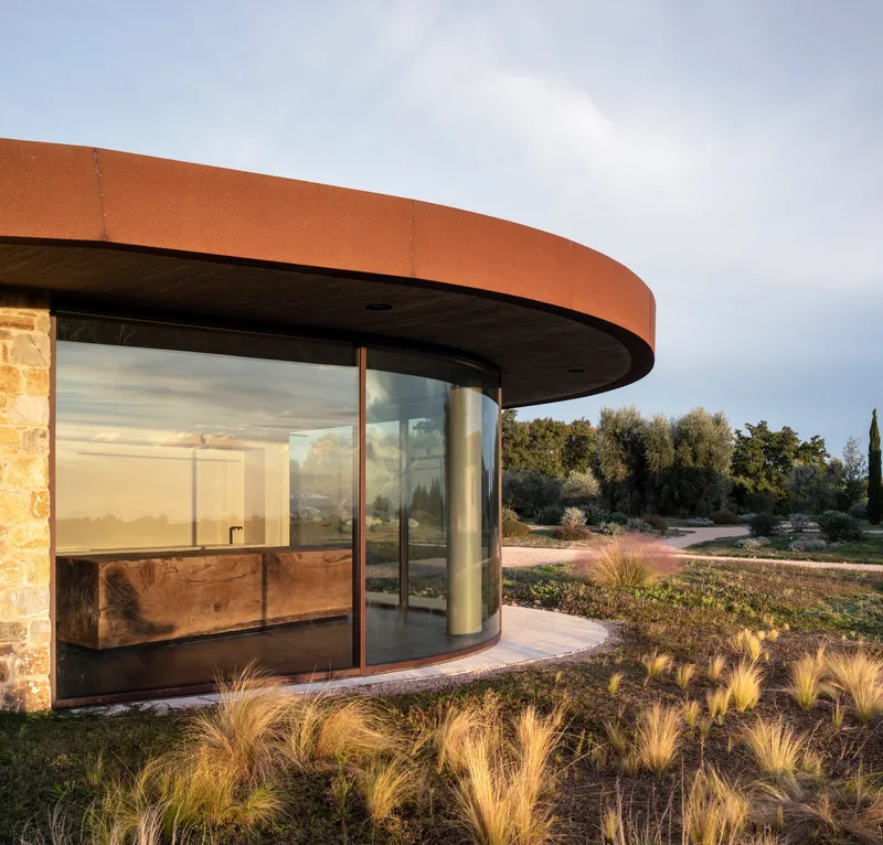 Curved glass facade with corten steel canopy and stone wall flanked by native grasses at dusk