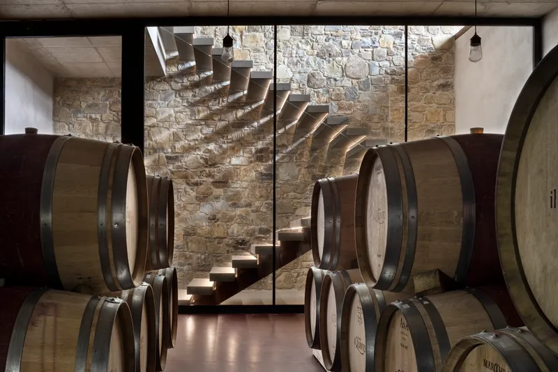 Cantilevered concrete staircase descending past stone walls and oak wine barrels
