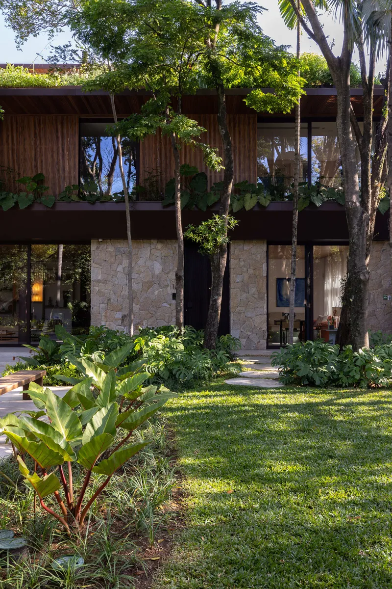 Two-story facade with stone base and timber cladding among mature trees and garden beds