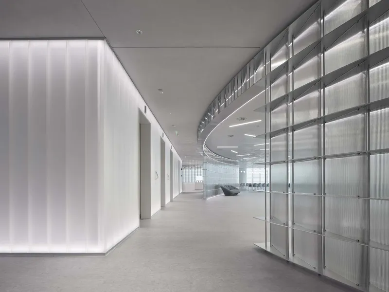 Curved corridor with translucent channel glass wall and backlit vertical panels along gray carpet floor