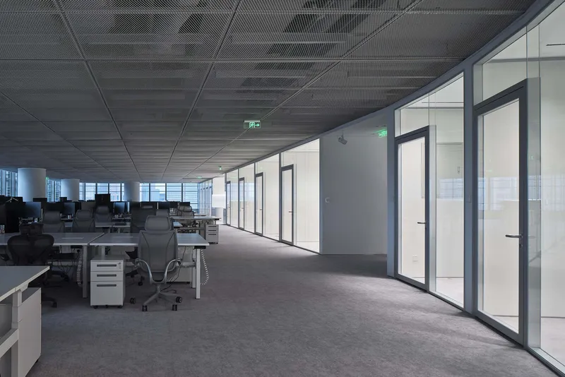 Open office floor with dark perforated ceiling panels and a corridor lined with glass-partitioned meeting rooms