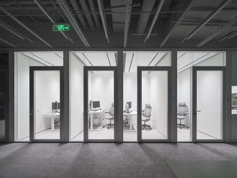 Four glass-walled office pods viewed from a corridor with exposed black ceiling and emergency exit sign