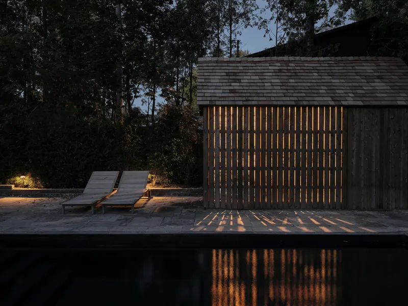 Vertical timber screen with interior light casting striped shadows on the pool deck at dusk