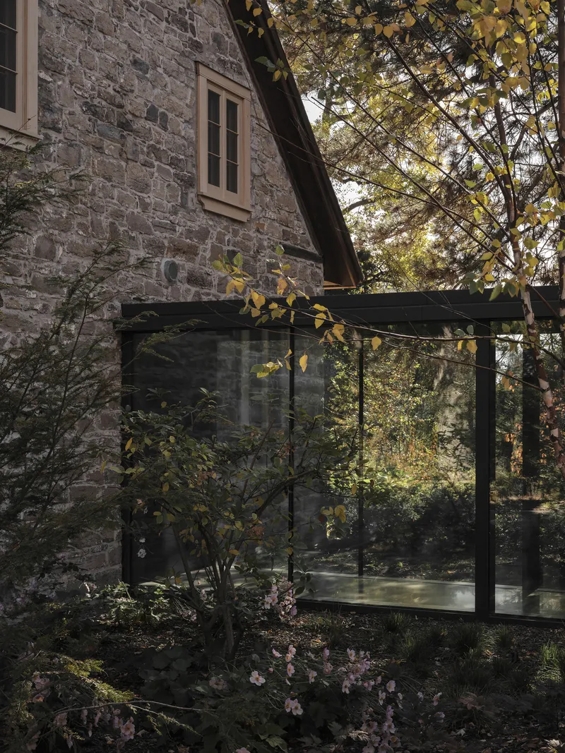 Glass pavilion addition adjoining the stone masonry wall with flowering plants in dappled sunlight
