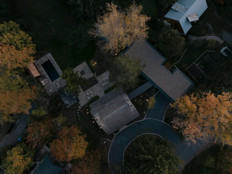 Drone view of stone and glass volumes with curved gravel driveway among autumn trees at dusk