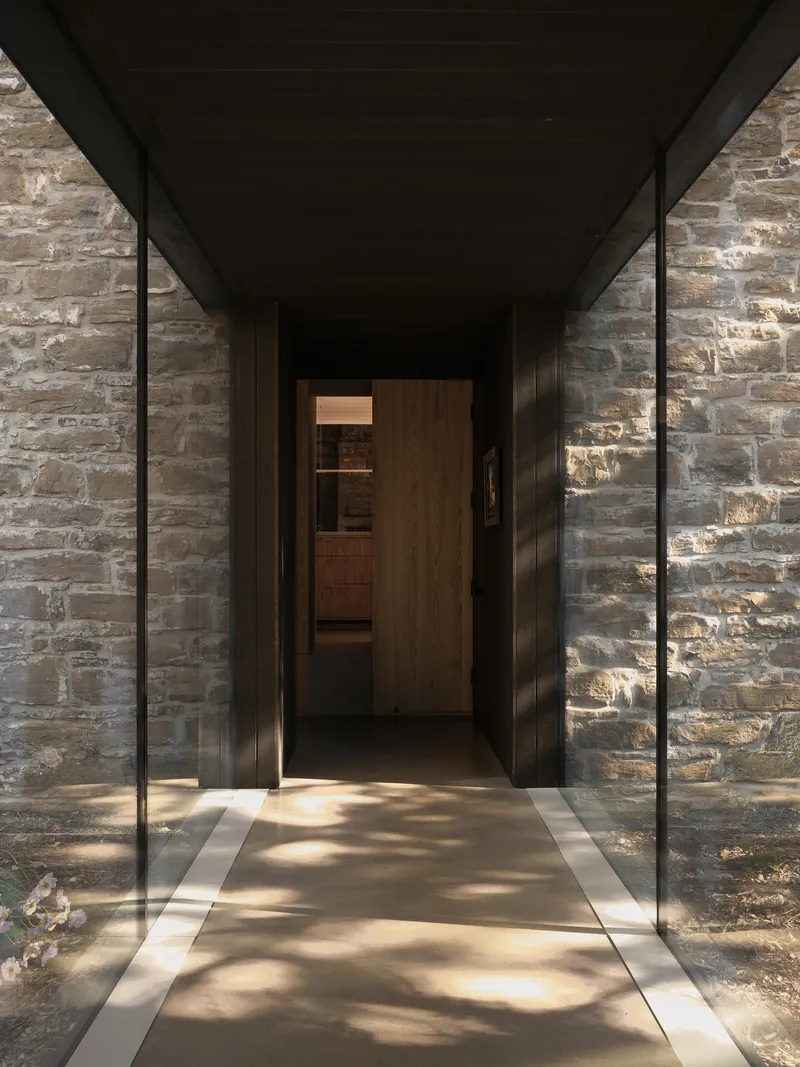 Covered entry passage with steel-framed glazing flanking timber doors and stone walls in dappled sunlight