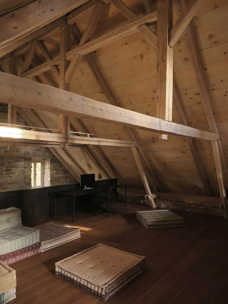 Attic bedroom with exposed timber roof trusses and planking above a dark workspace and floor cushions