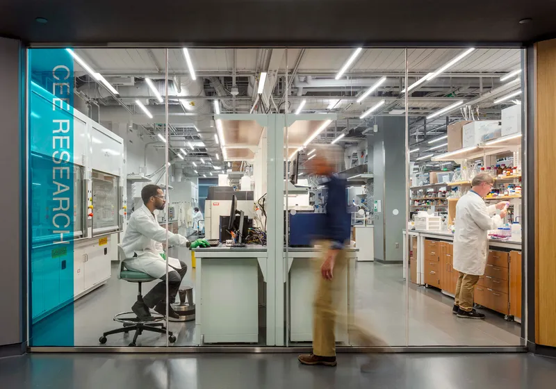 Glass-walled research laboratory with exposed ceilings, lab benches, and researchers in white coats