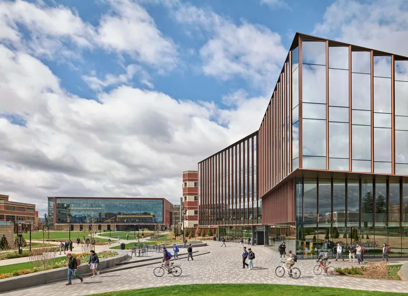 Campus plaza with paved walkways and planted beds linking vertically striped facade volumes under daytime clouds