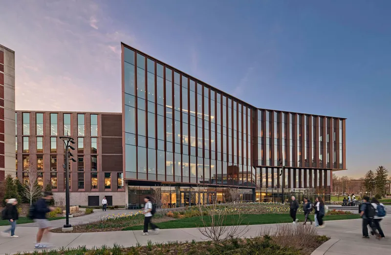 Glass and metal vertical fin facade with ground-level entry and pedestrians crossing lawns at dusk