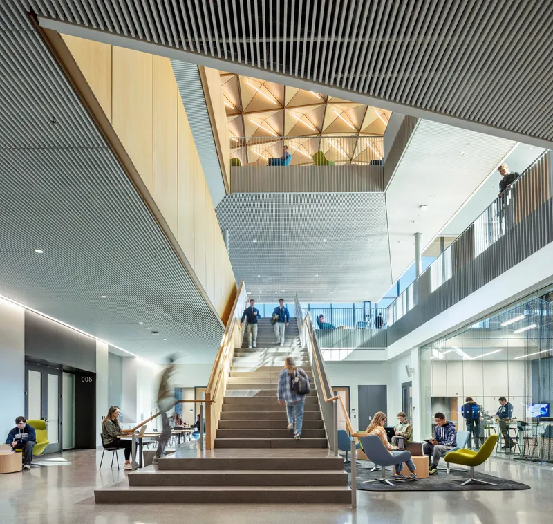 Multi-story atrium with wide central staircase and triangulated wood ceiling panels above seated students