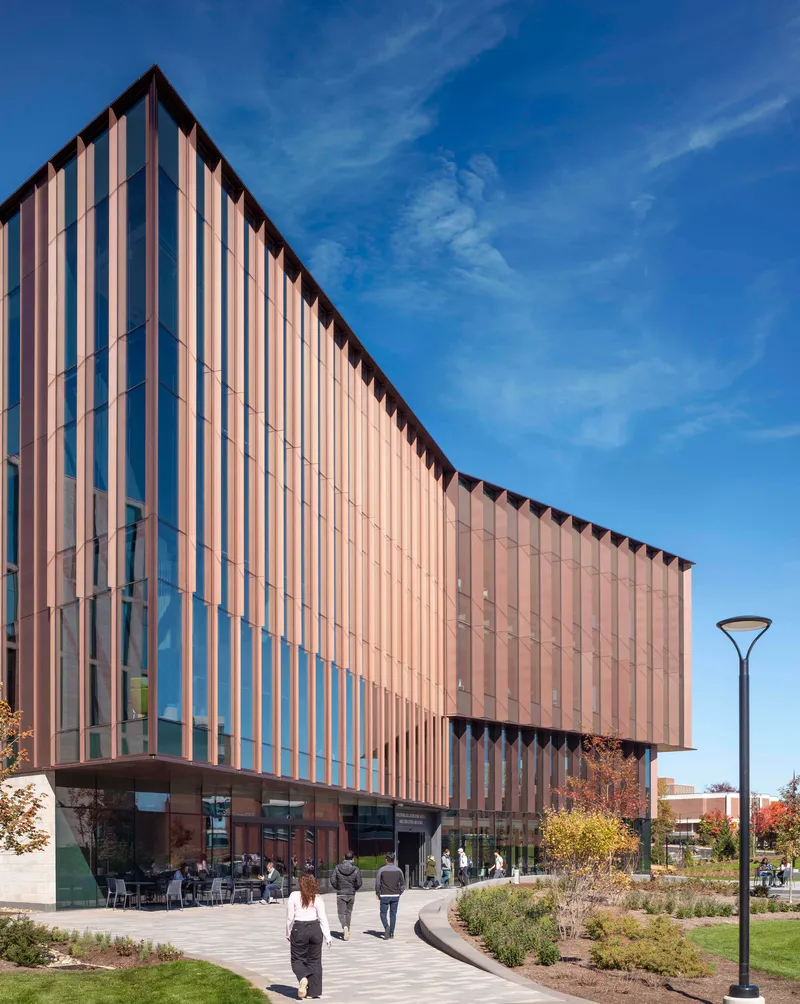 Curved facade with vertical metal fins rising above landscaped plaza in afternoon light