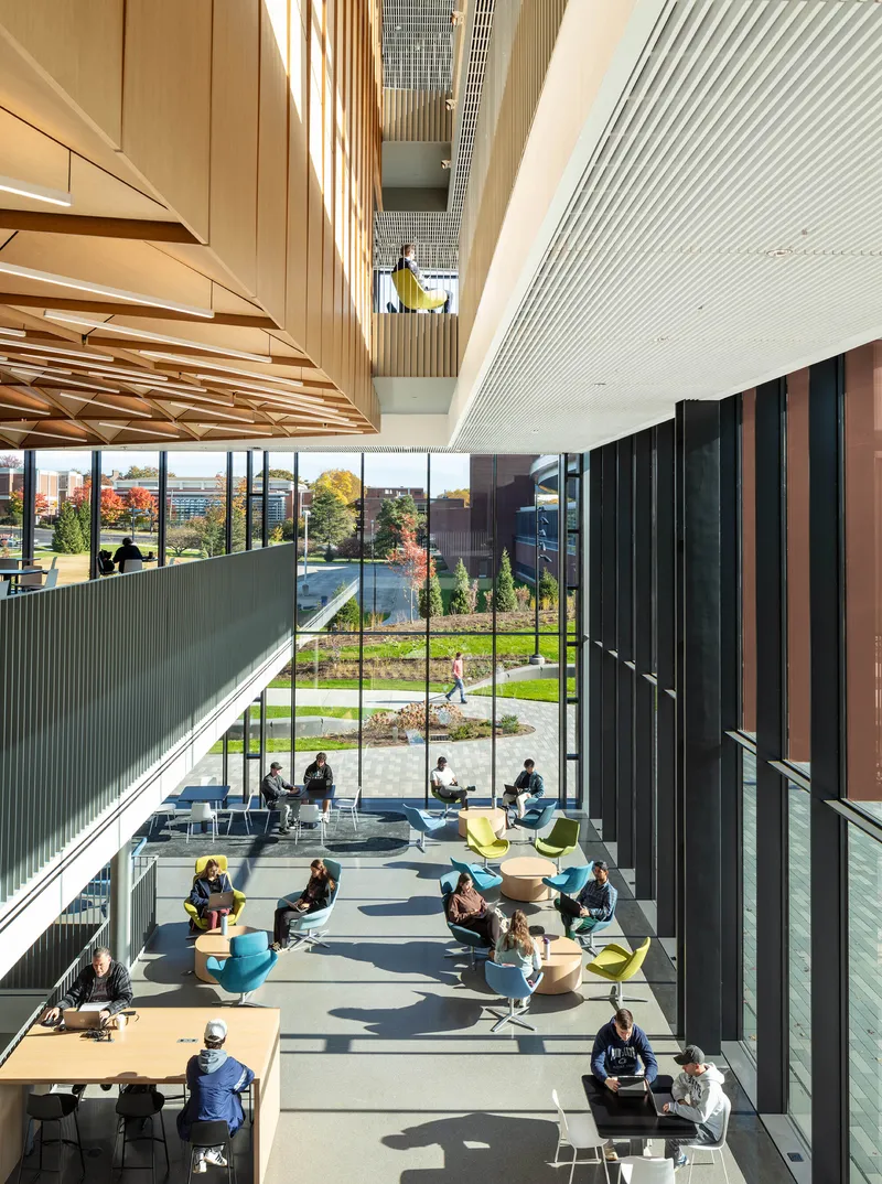 Atrium lounge with scattered colorful seating, floor-to-ceiling glazing overlooking the campus, and triangulated wood ceiling above