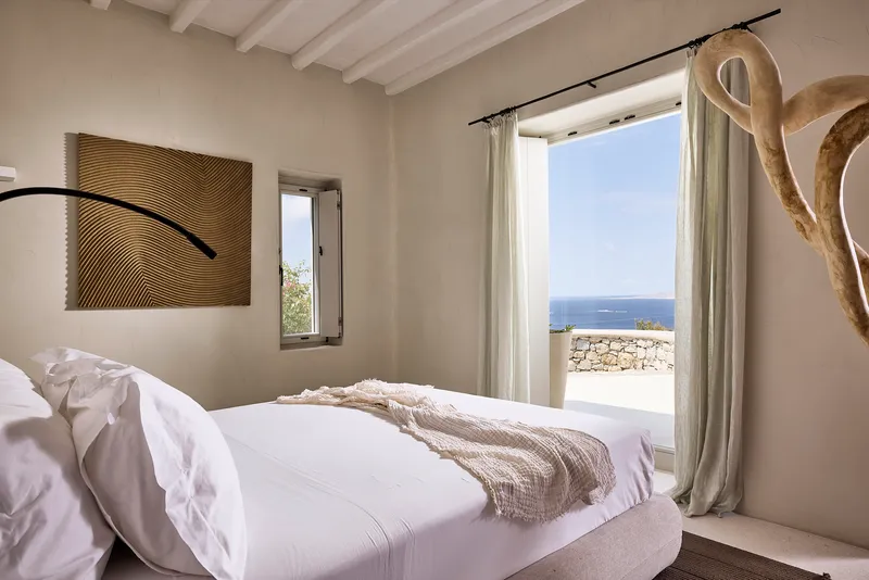 Bedroom window framing ocean view with stone wall terrace and linen curtains under blue sky