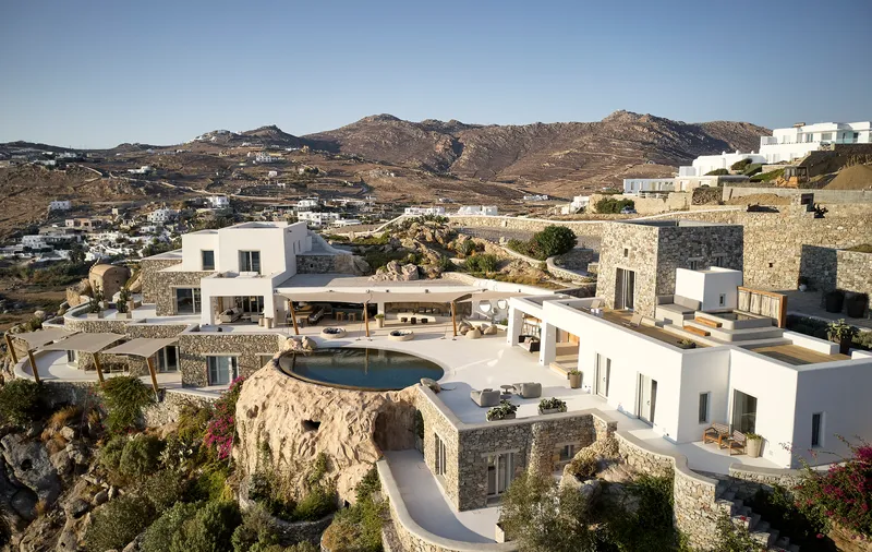 Aerial view of white plaster volumes and stone-clad structures with circular pool terrace on rocky hillside terrain