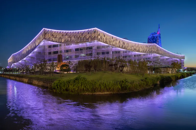 Illuminated undulating facade with purple lighting reflected in the adjacent waterway at dusk