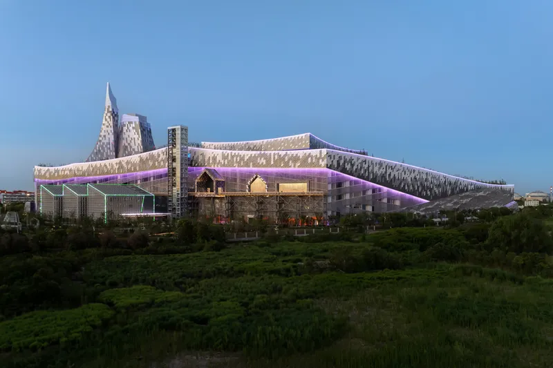 Illuminated perforated facade with purple accent lighting viewed across a green field at dusk