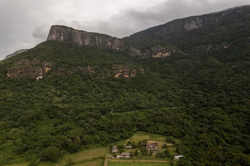 Aerial view of a small compound clearing in forested terrain with dramatic rock escarpments rising behind