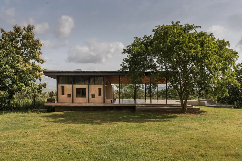 Single-story pavilion with deep overhanging roof, steel columns, and glazed walls framed by mature trees under a partly cloudy sky