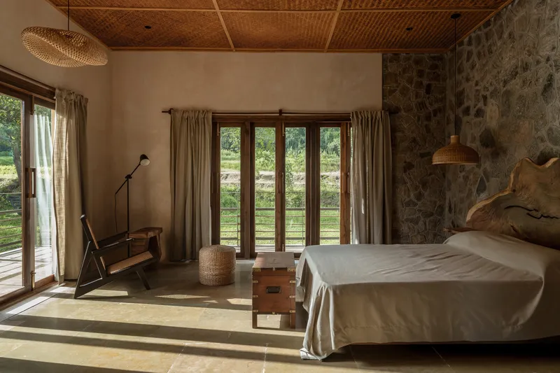 Bedroom with woven bamboo ceiling, stone accent wall, and morning sunlight streaming through timber French doors